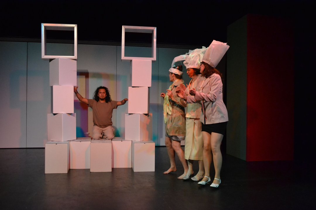 Drei Frauen mit Papierhüten stehen rechts auf der Bühne und schauen auf einen Jungen,  der einen Turm aus weißen Pappelementen gebaut hat und festhält