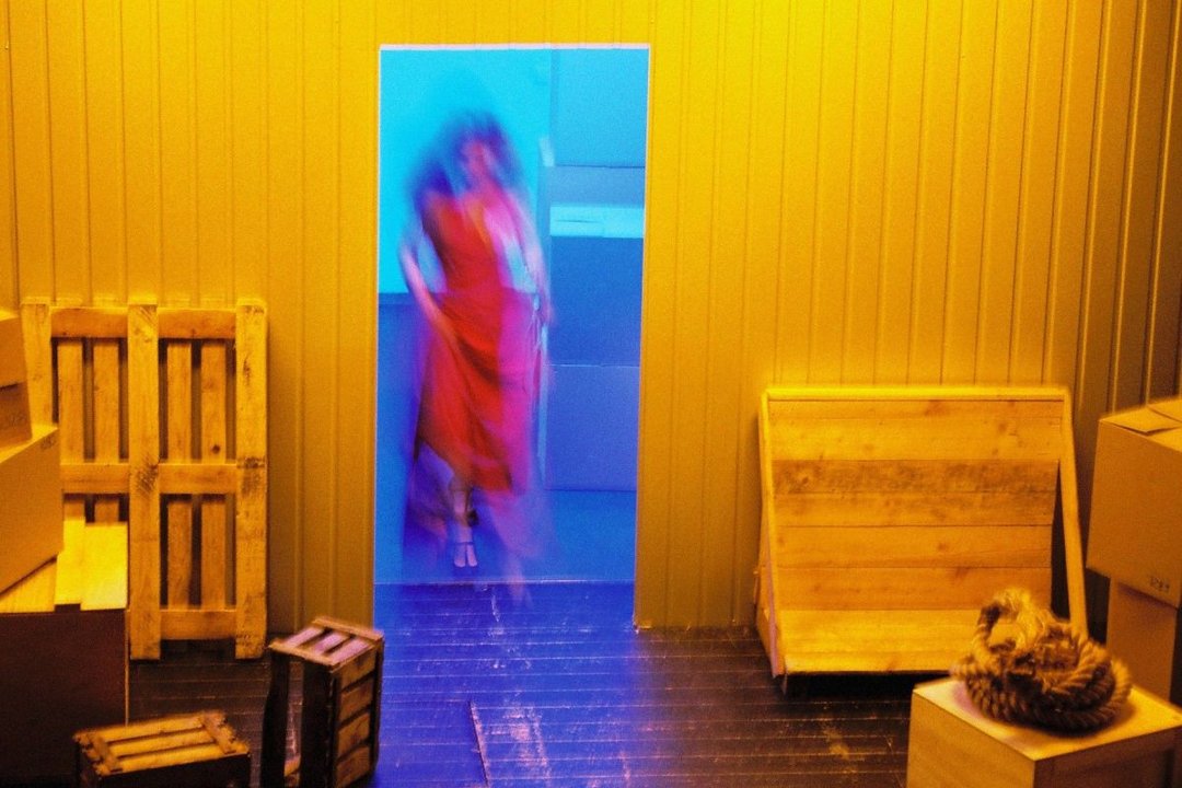 Bühnenbild Frachtraum, Tür in blauem Licht, an der Tür steht eine Frau in einem roten Kleid.