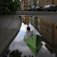Frau sitzt in Papierschiffchen auf einer Straßenpfütze, in der sich die Häuser an der Straße spiegeln